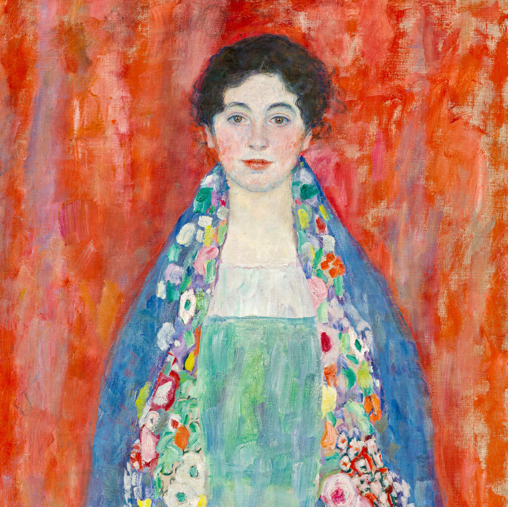 A portrait of a young woman by Gustav Klimt. She is dressed in an ornate dress and multicolored robe, staring out at the viewer, against a bright red background.