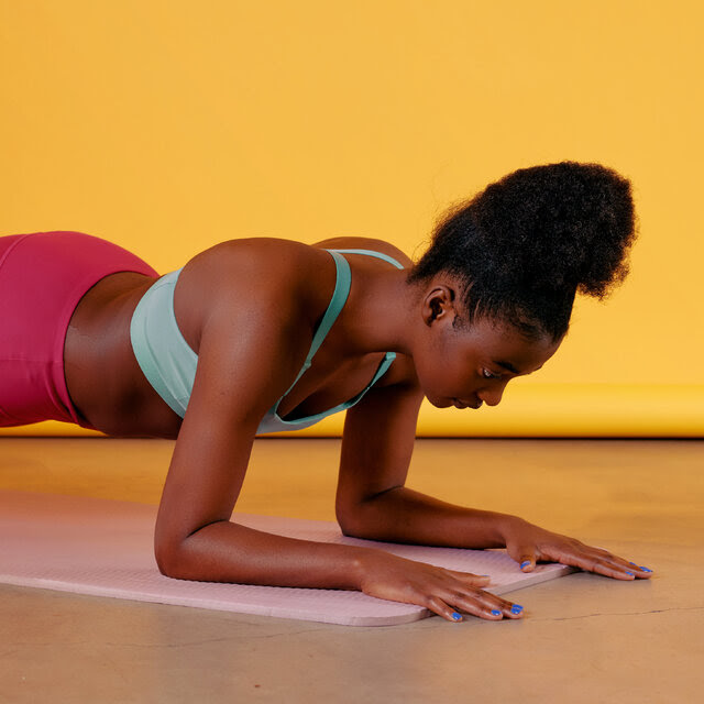 A woman is holding a plank position on a pink yoga mat. She is wearing a light blue sports bra and dark pink leggings and has her hair up in a high ponytail.
