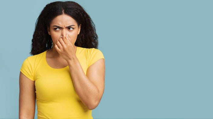 Studio portrait of a young woman holding her nose like something smells bad. She is wearing a yellow t-shirt on a light blue background.