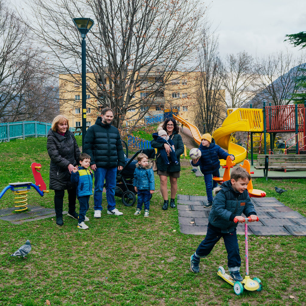 The Baldo family in a playground. An apartment building, trees and mountains are visible behind them.