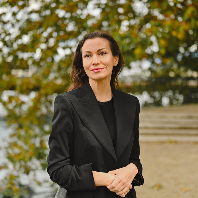 Irina Bolgar wearing a black suit in front of trees with her hands crossed in front of her. 