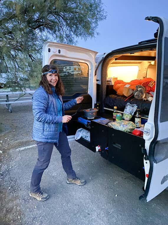 Una persona parada junto a una camioneta mientras cocina con utensilios de cocina portátiles.