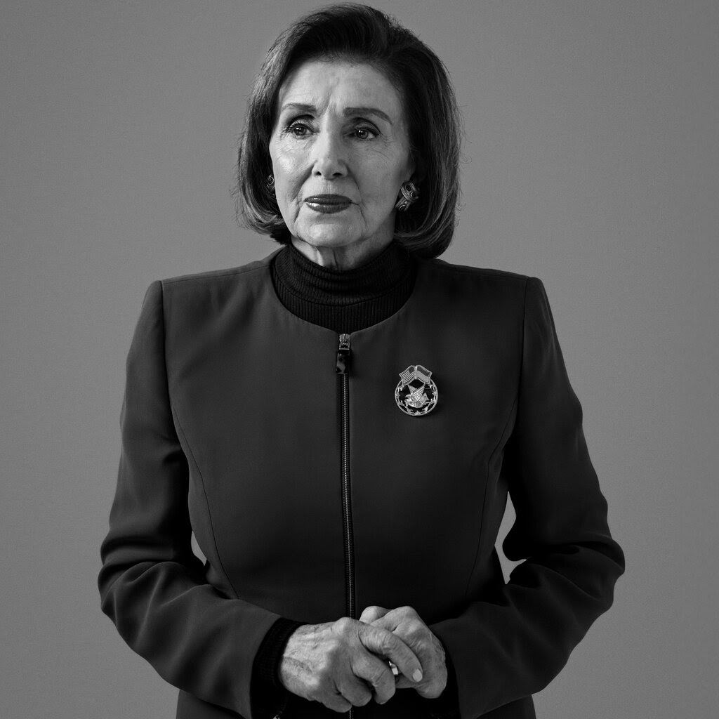 A black-and-white portrait of Nancy Pelosi, looking pensive, with an American eagle broach on her zipped collarless jacket.