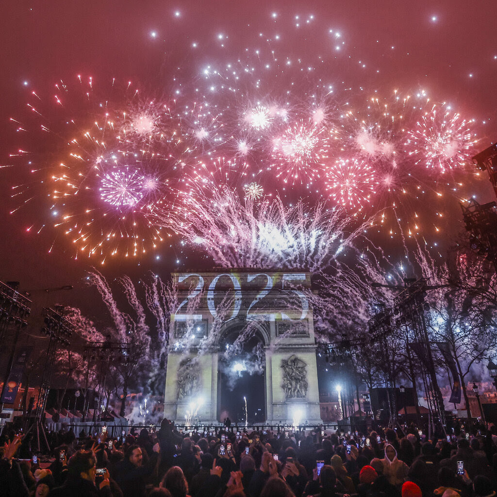 People look up at red and orange fireworks in the sky. The number “2025” is projected on a large arch nearby.