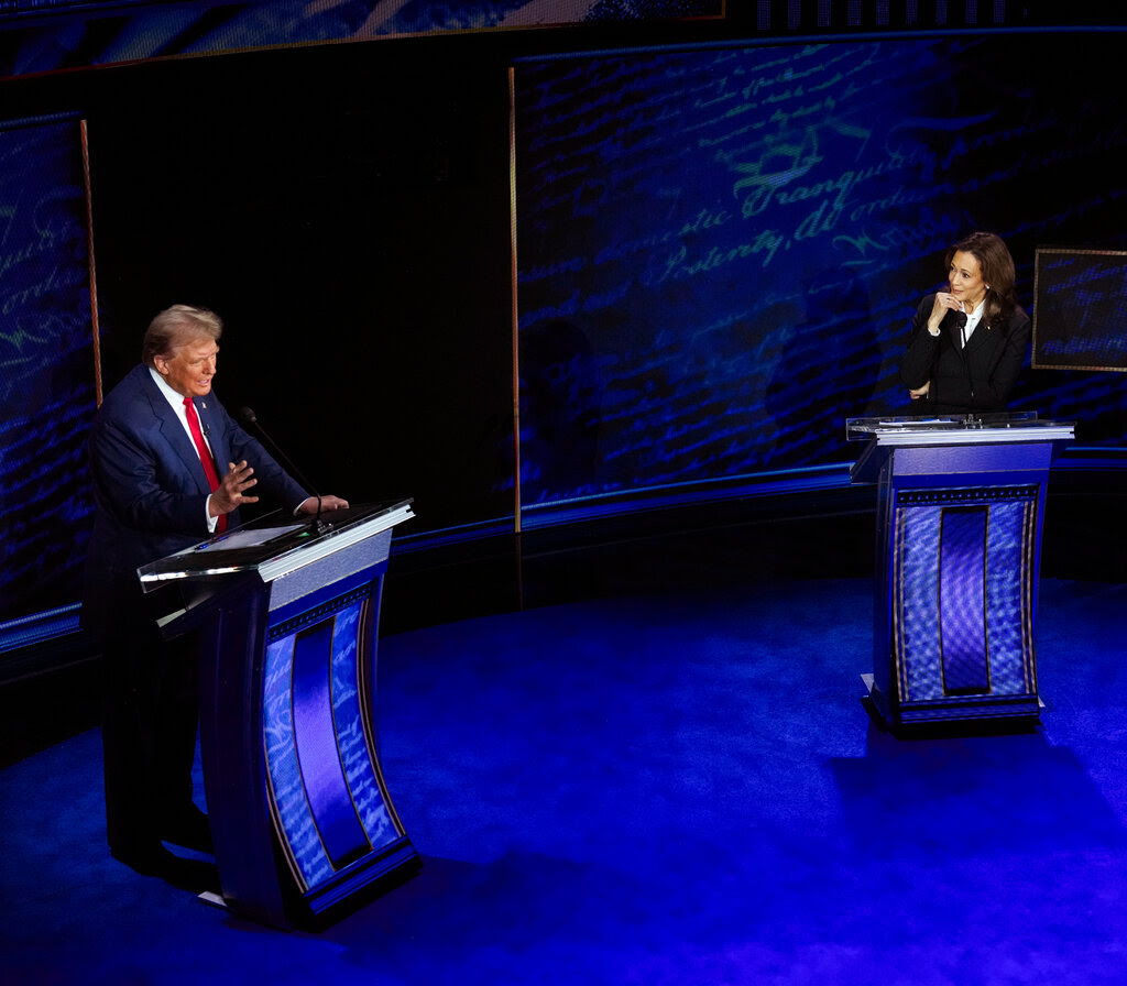 Kamala Harris and Donald Trump behind  lecterns. She is watching him speak, her head tilted and a hand under her chin.