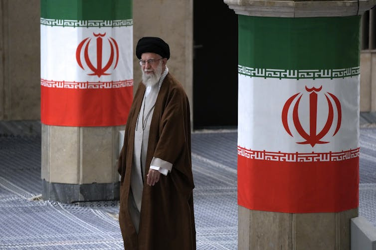 A bearded man in clerical robes walks between two pillars festooned with green, white and red flags.