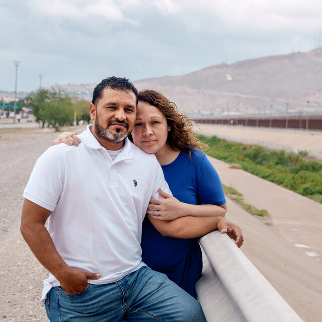 Sandra Reyes stands behind her husband, Hector Reyes, with one hand on his shoulder and the other on his arm, on a roadside with the U.S.-Mexico border behind them.