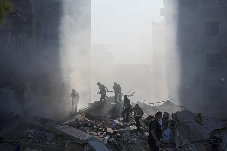 A team of people walk up a pile of debris and rubble holding a hose to put a nearby fire out.