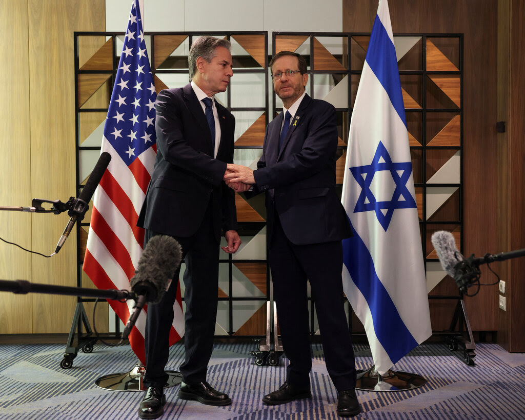 Antony Blinken and Isaac Herzog, wearing suits, stand in front of their country’s flags, shaking hands.