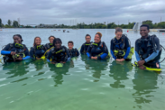 Nine people pose in waist deep water all dressed in scuba gear as they learn how to dive. 