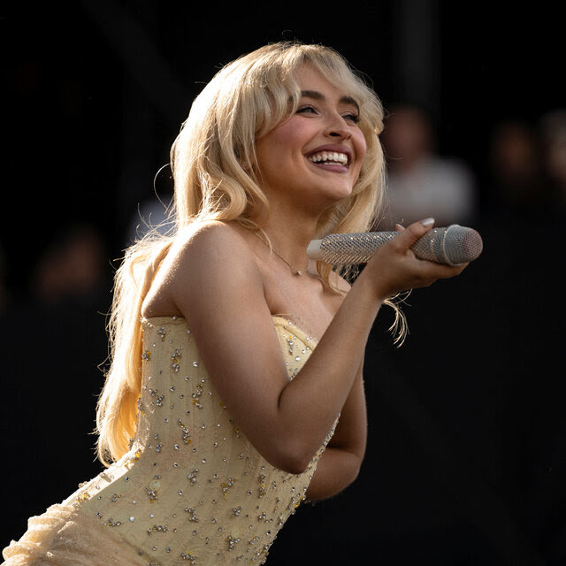 A blond woman in a yellow sequined strapless top smiles and holds a microphone out toward a presumed audience.