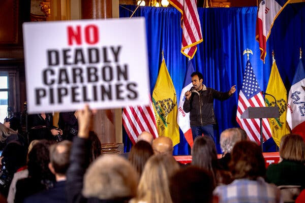 Vivek Ramaswamy speaking on a stage with  roughly 20 people looking on. A person in the foreground is holding up a sign that says No Deadly Carbon Pipelines.