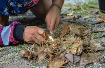 A pair of hands work to start a spark from flint and steel, flame leaping to life in the dry tinder of a survival fire.