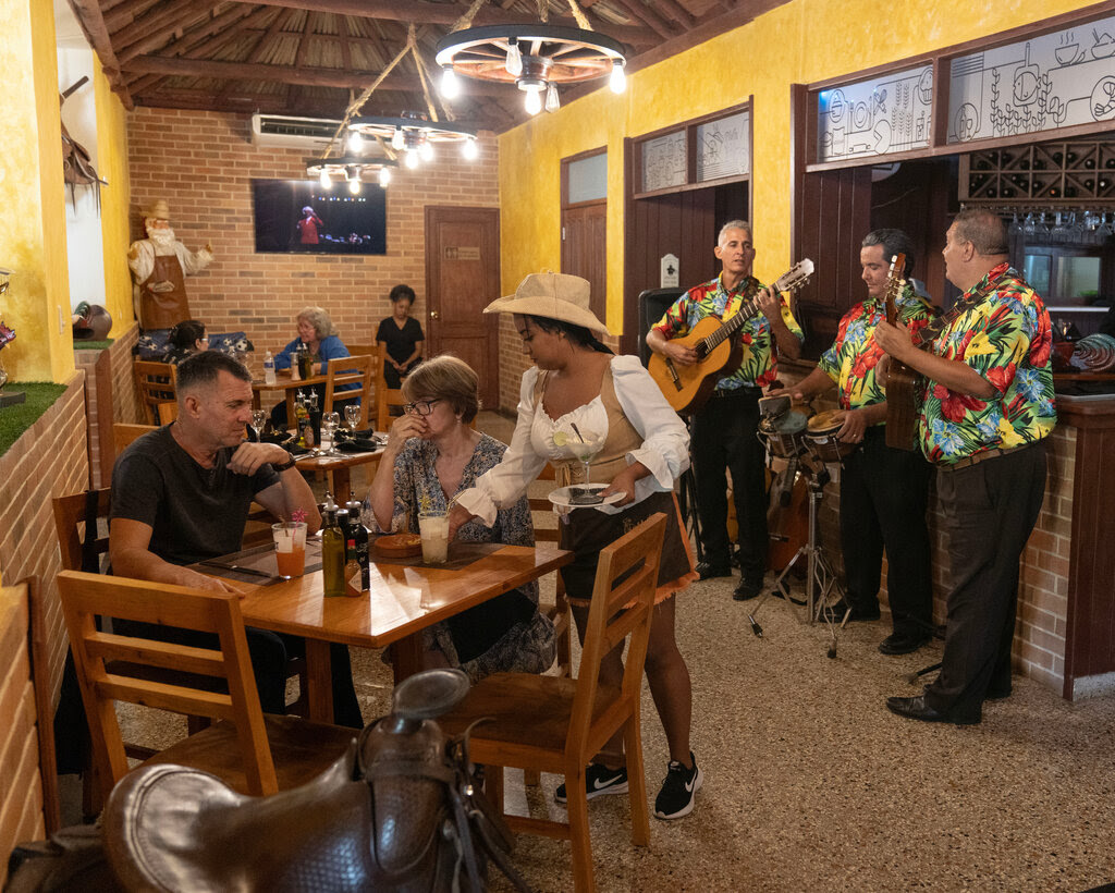 A scene at a restaurant as a woman serves two people at a table and a trio with two guitarists and a drummer plays music.