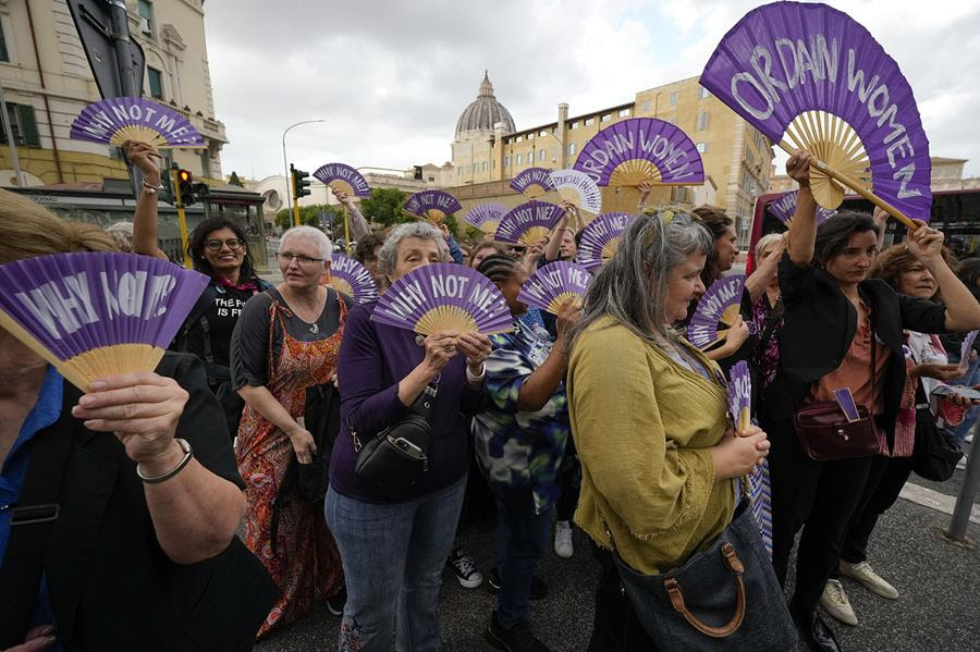 Women holding purple fans that say "Ordain women" or "Why not me?"