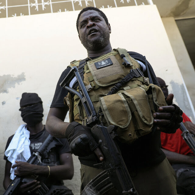 A man wearing a military-style vest and holding a rifle stands just in front of other men, some of whom are wearing masks over their faces.