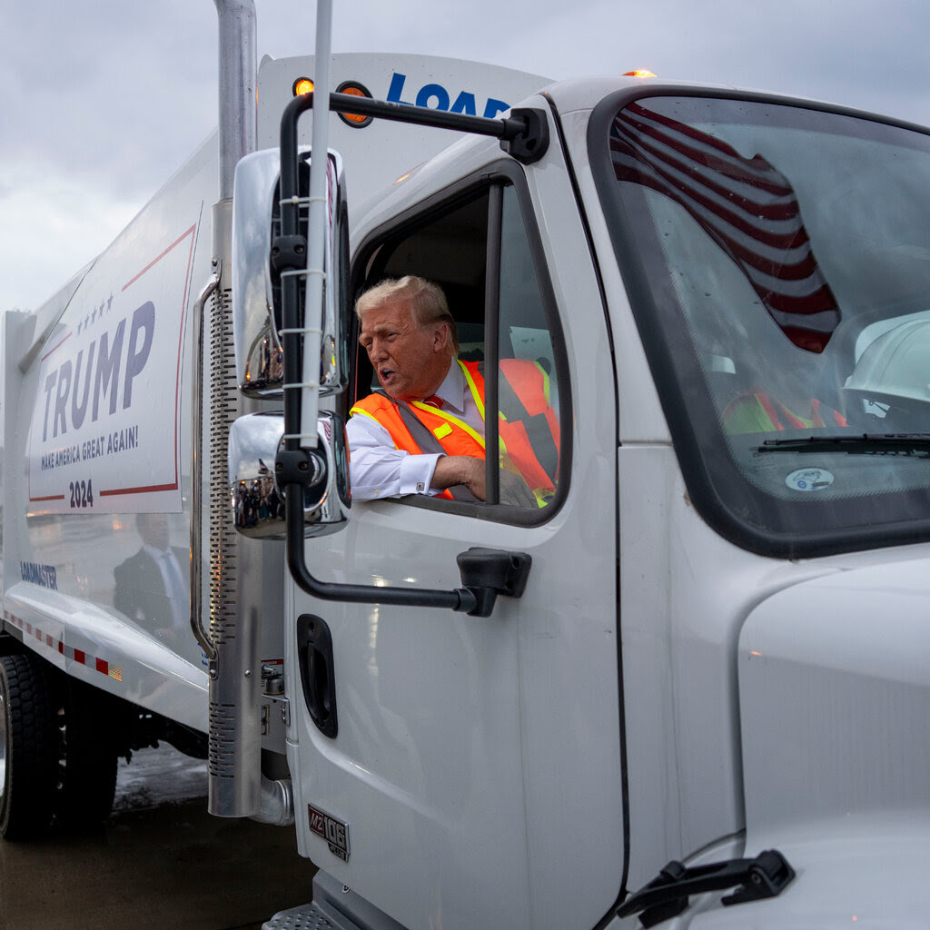 Donald Trump in a hi-viz orange vest rides in a truck cab.