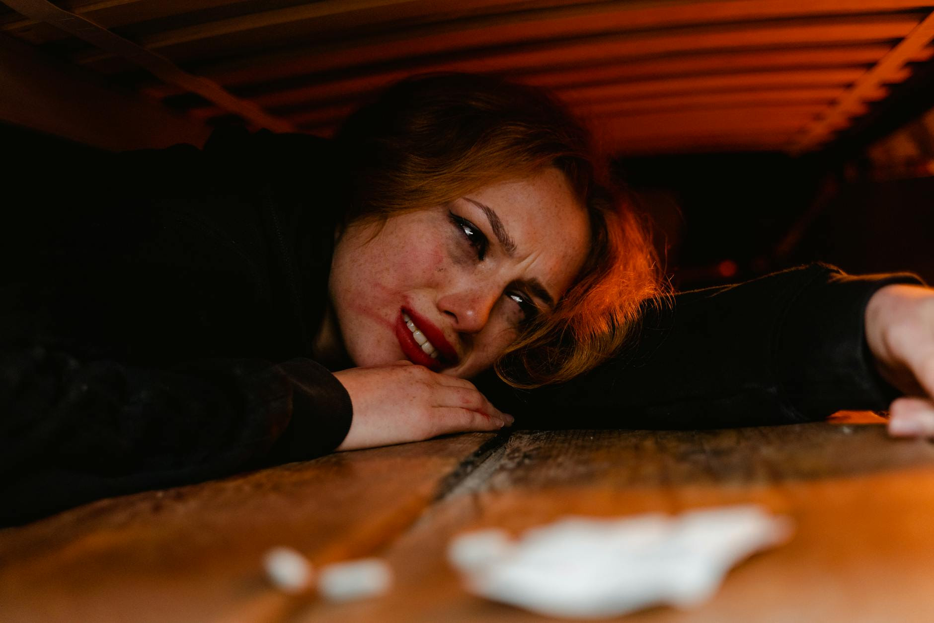 A distressed woman lying under a wooden structure, expressing fear and anxiety.