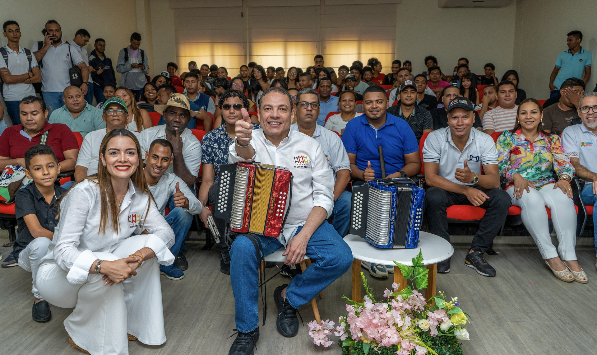 Un grupo de personas sonríe en un salón de eventos. En el centro, un hombre toca un acordeón mientras muestra un gesto de aprobación con el pulgar levantado. A su alrededor, hay otras personas que también están sonrientes y sentadas en varias filas, mostrando un ambiente alegre y participativo.