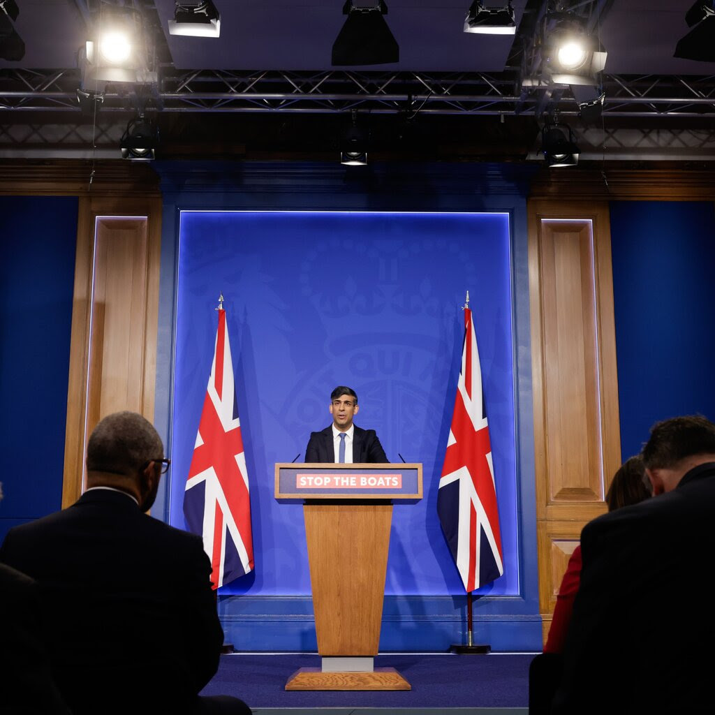 Prime Minister Rishi Sunak of Britain standing between two British flags at a lectern with a sign that says “Stop the Boats” on it.
