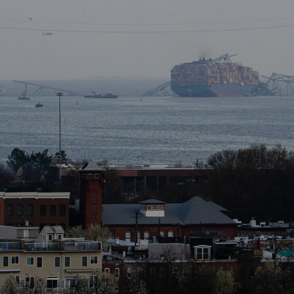 A number of buildings in the foreground, and in the background is a river and the remains of a bridge with the massive cargo ship that ran into it.
