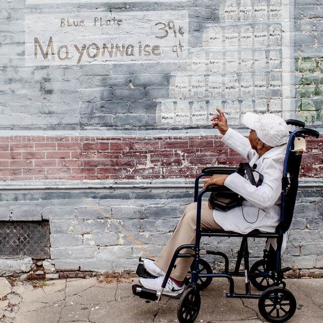 Mamie Kirkland in tan slacks and a whiteish sweater and baseball cap sits in a wheelchair and points up at a mural on a brick wall.