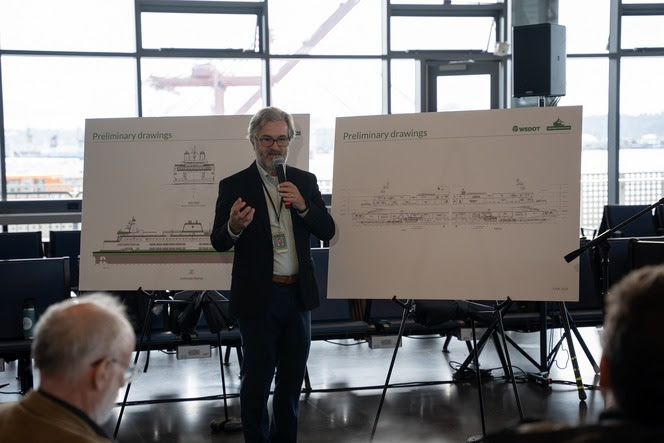 Person holding microphone in front of poster boards with ferry renderings speaking to people seated