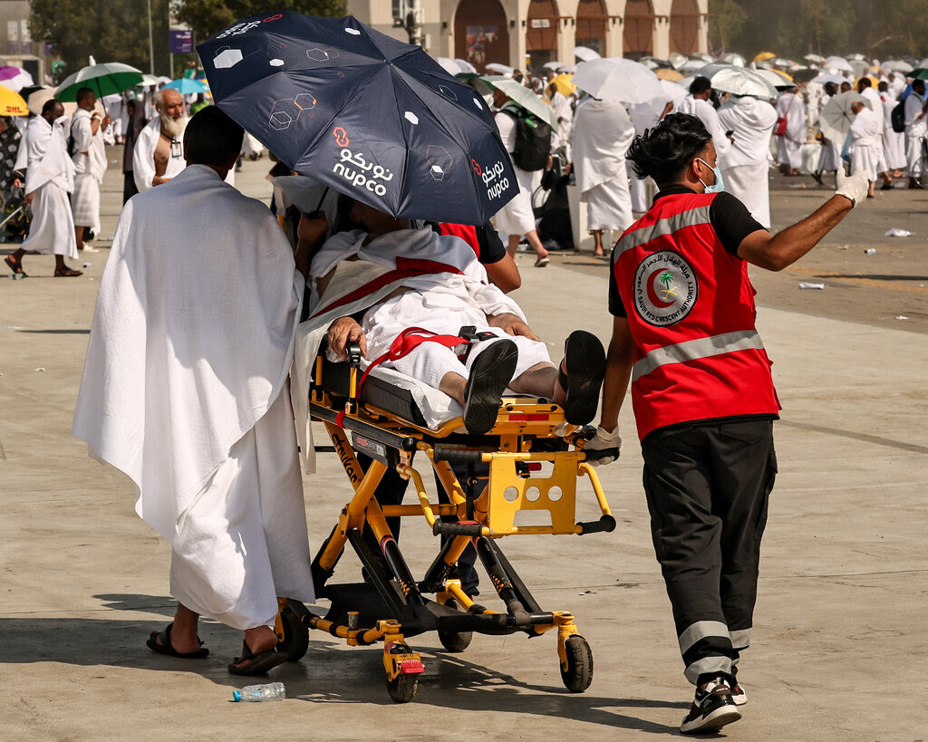 A person is wheeled in a stretcher, with a dark umbrella covering the face. Pilgrims in white clothing stand with umbrellas in the background.