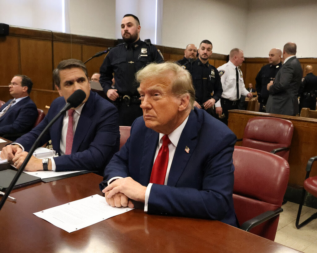 Former President Donald J. Trump sitting at a table. He is in a dark suit. He has his hands on paper.