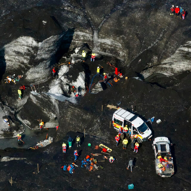 An aerial view of rescue personnel and emergency vehicles dotting an Icelandic glacier, which is a blend of black and white. 