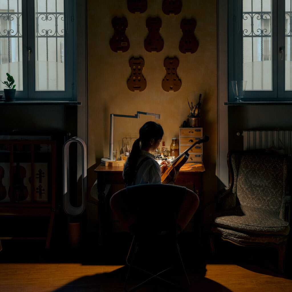 A silhouette of the violin maker is seen at a table in the darkened studio. She meticulously adds details to make the instrument look more antique.