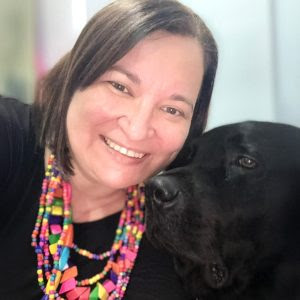 A imagem mostra a foto de Karem, uma mulher sorridente ao lado de um co preto. Ela tem cabelo castanho escuro e usa um colar colorido