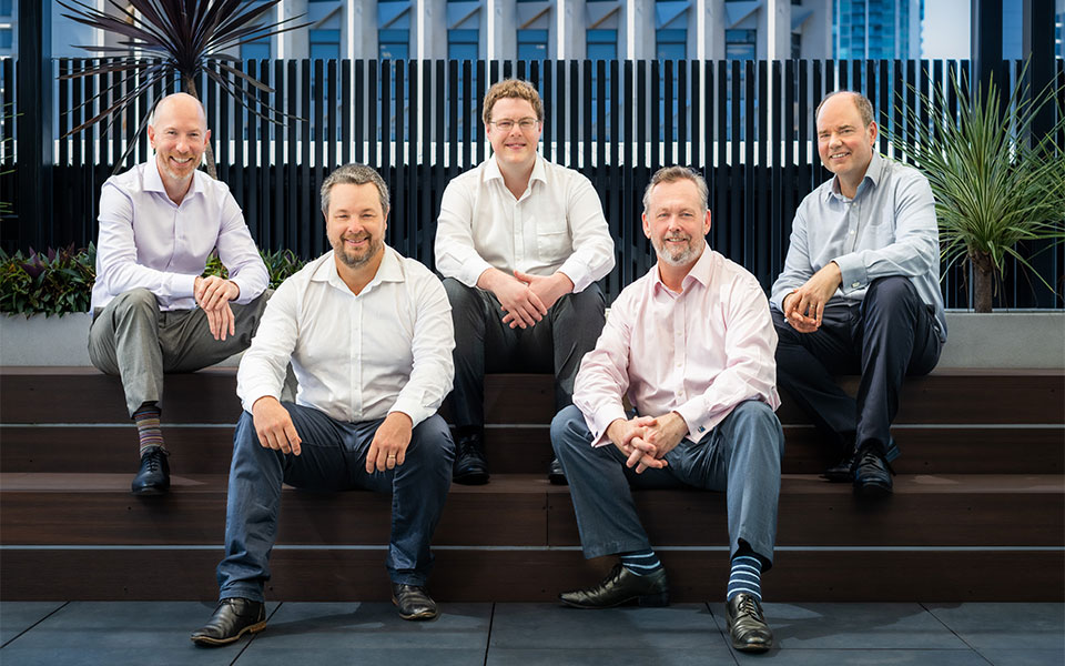 The image shows five men in business casual attire, smiling and seated casually on steps in what appears to be an outdoor section of a modern office building, suggesting a relaxed yet professional team environment.