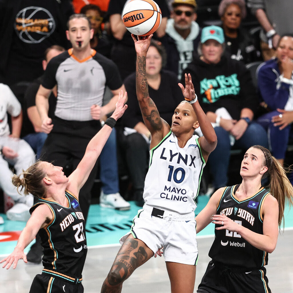 A Lynx player in a white jersey jumps and shoots, with Liberty players in black jerseys on either side of her.