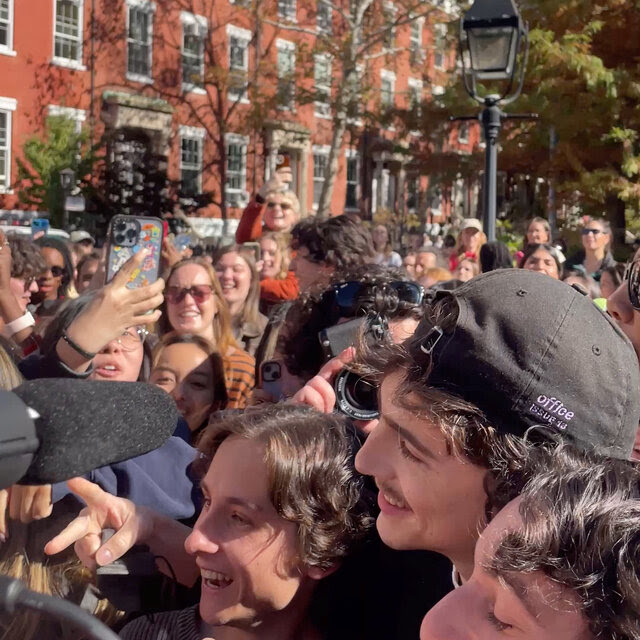 Timothée Chalamet poses for a photo with two look-alike fans. A crowd surrounds them, some taking pictures.