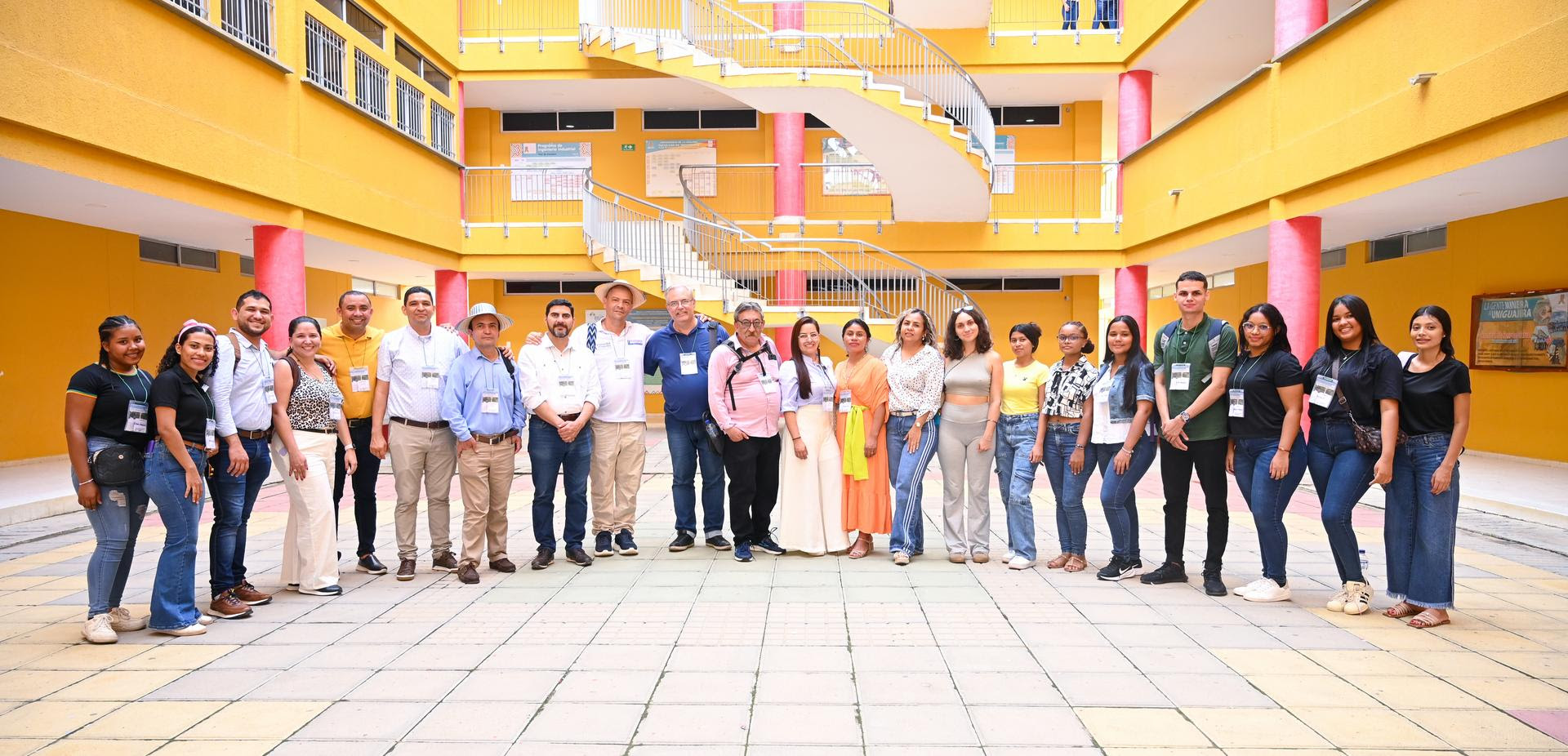 Un grupo de personas posando para una foto en el patio de un edificio de varios niveles, de colores amarillos y rosados. Hay una mezcla de hombres y mujeres en la imagen, algunos vestidos de manera casual, mientras que otros visten ropa más formal. Todos llevan gafetes de identificación colgados alrededor del cuello.