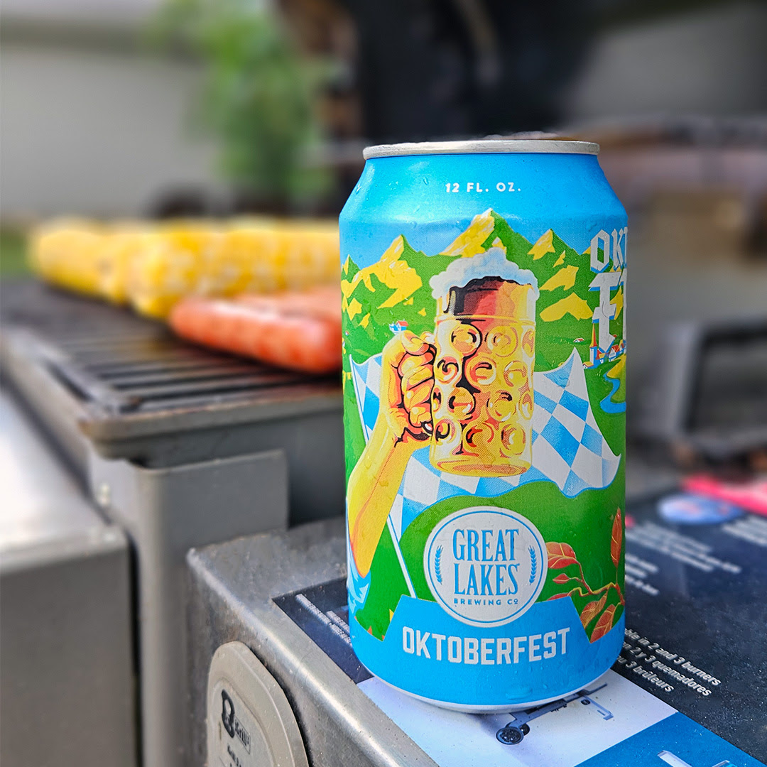 12 oz. Can Oktoberfest on the side of a grill, with hot dogs and corn on the grill.