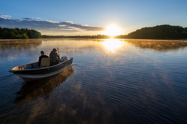 two people with fishing gear in a boat, as the reflection of a blazing yellow sunrise reflects along the still surface of the large lake