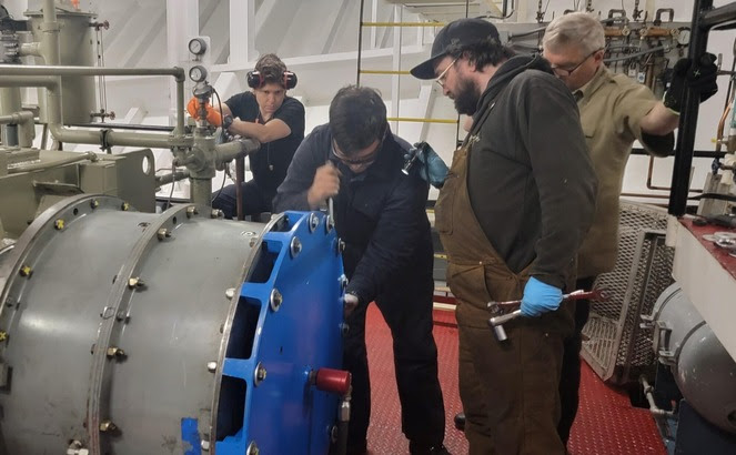 Four people working on faulty propulsion clutch in the engine room of a ferry