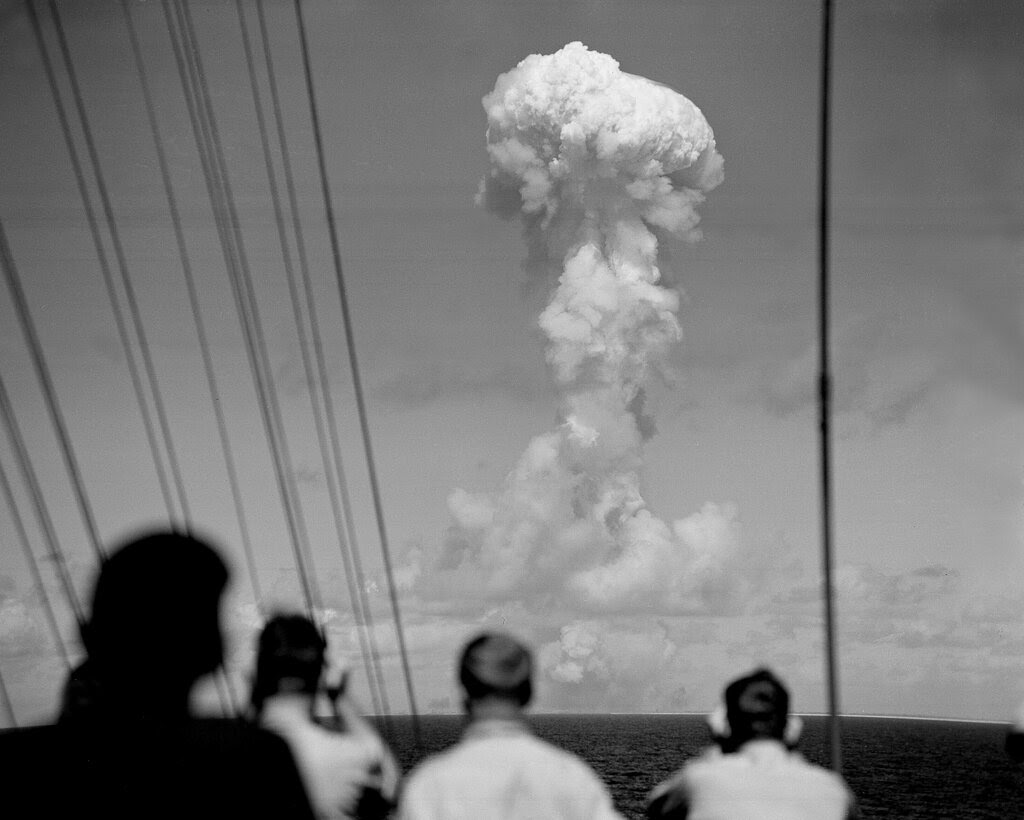 Four people standing on the deck of a ship face a large, white mushroom cloud in the distance. 