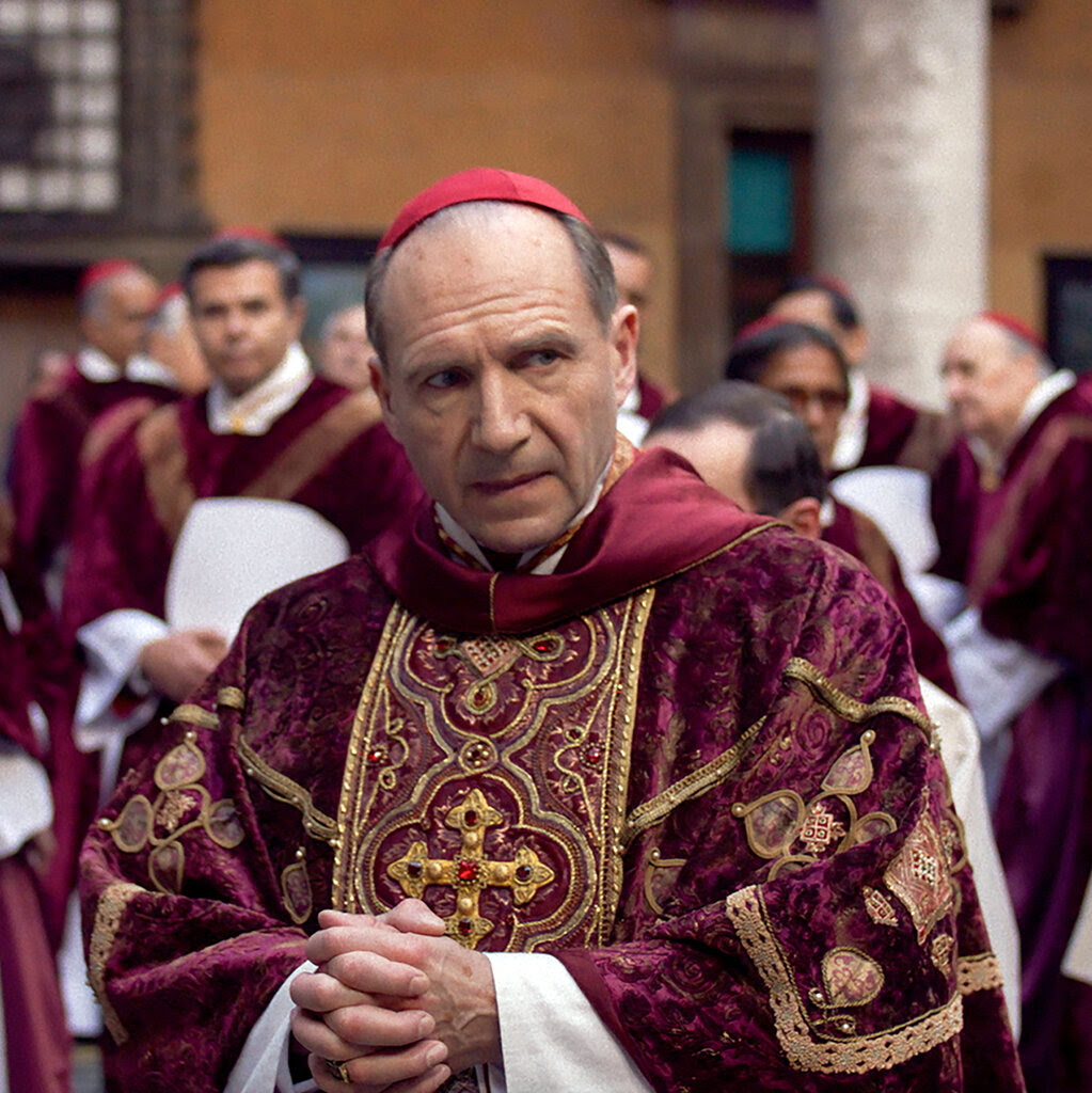 In a movie scene, Ralph Fiennes, wearing a red skullcap and an ornate magenta robe, looks concerned. His hands are clasped, and he’s surrounded by other figures in robes similar to his. 