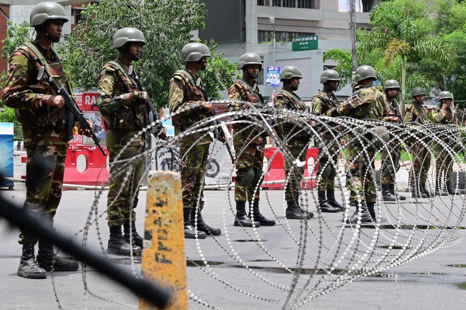 Bangladeshi army personnel stand in a line behind barbed wire as they guard the parliament house in Dhaka, Bangladesh, following clashes between police and protesters. The soldiers wear green camouflage fatigues and helmets, and they hold rifles as they look straight ahead.