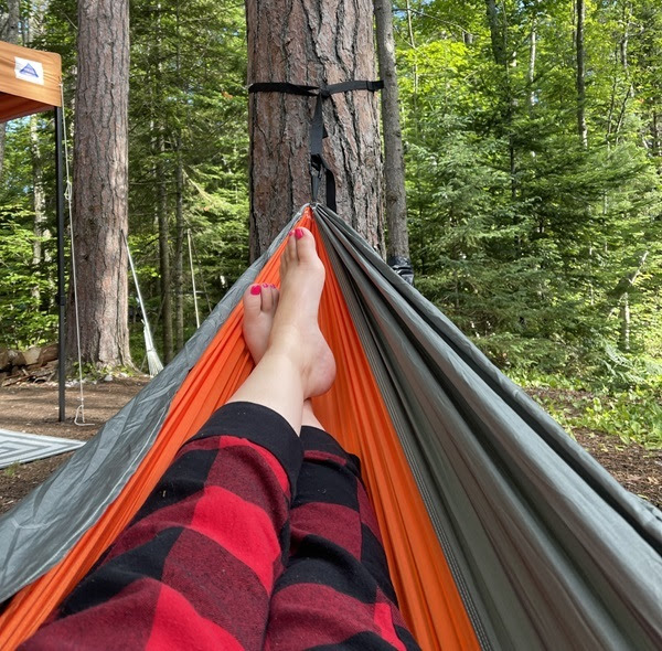 a woman's legs in red and black plaid sweats, with bare feet and pink-painted toes, stretched out in a orange and black tree hammock