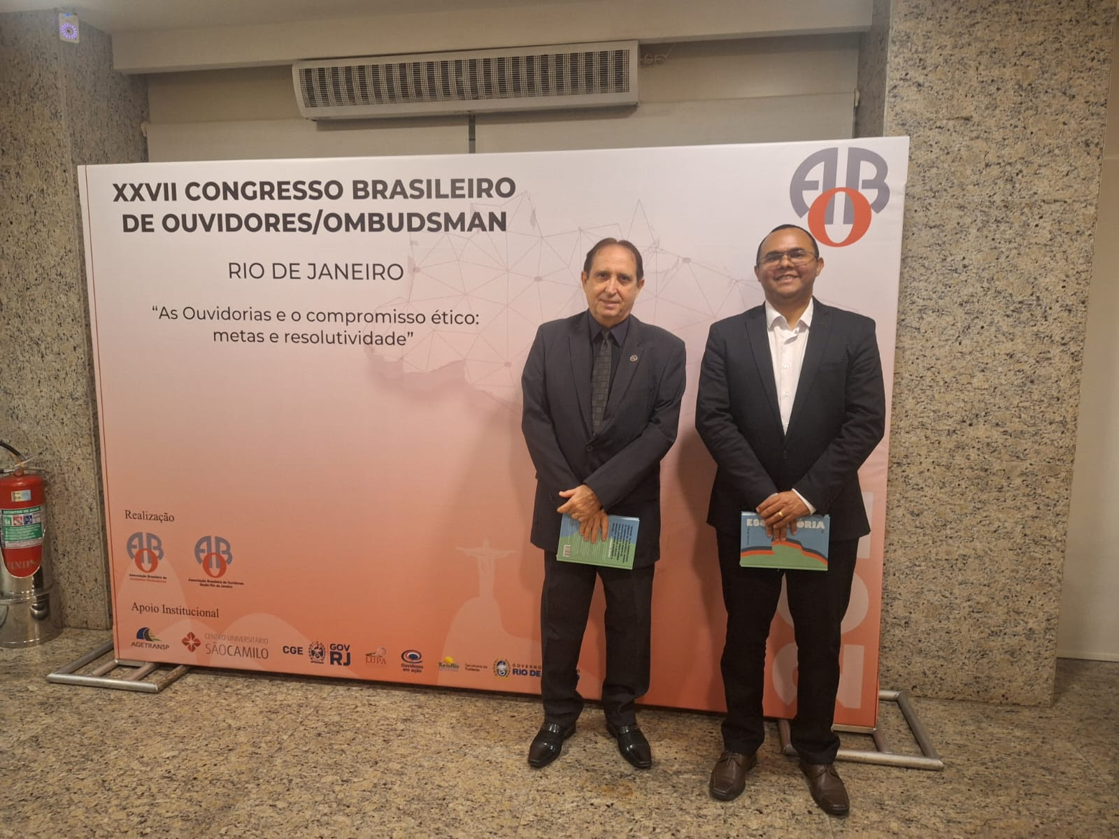 a imagem mostra, em primeiro plano, dois homens usando roupas sociais, ao fundo um painel nas cores branca e marrom exibem a mensagem "XXVII congresso de ouvidores do Brasil - Rio de Janeiro - As ouvidorias e o compromisso ético: metas e resolutividade¿, na parte inferior do painel são apresentadas logos das instituições realizadoras e parceiras do evento