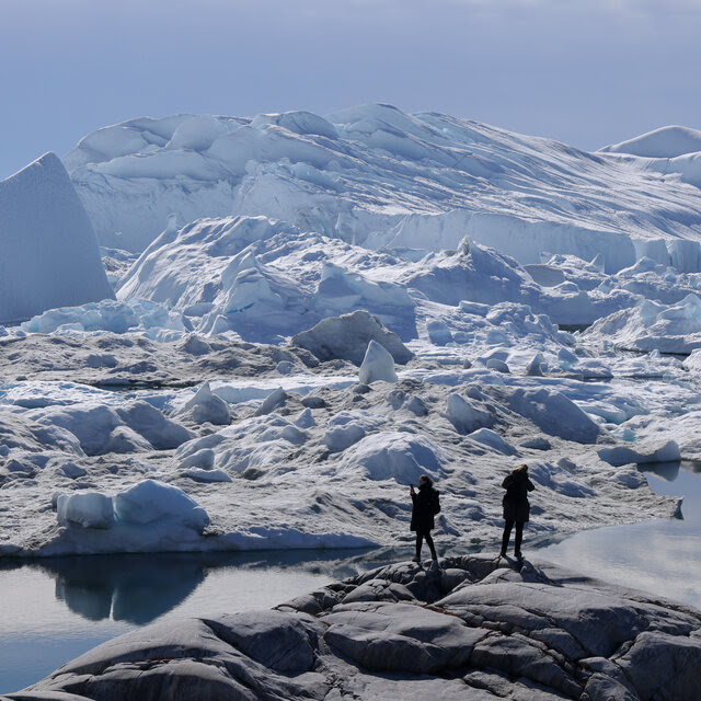 Two people stand in the foreground. Icebergs are in the background.