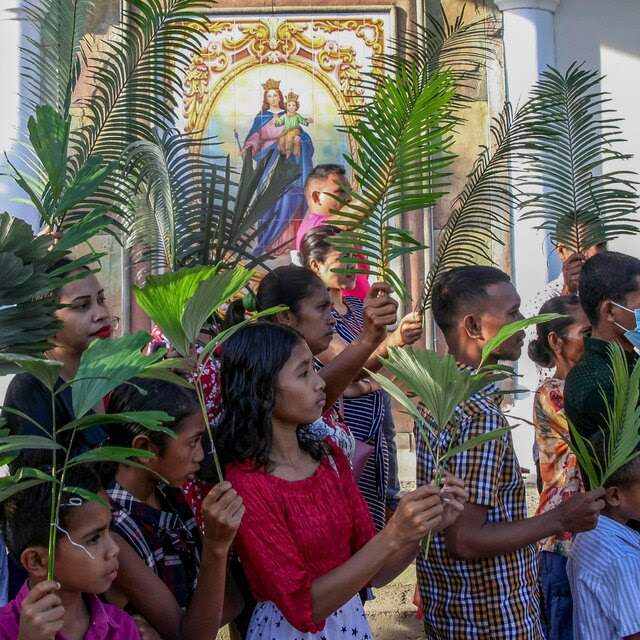 A line of people stands in front of a church, each person holding a palm frond.