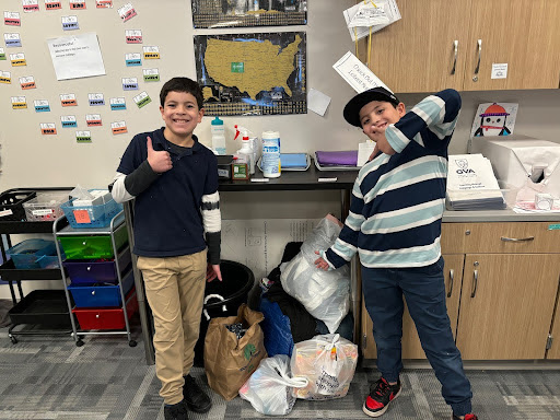 Two boys giving thumbs up next to bags of supplies in a classroom.
