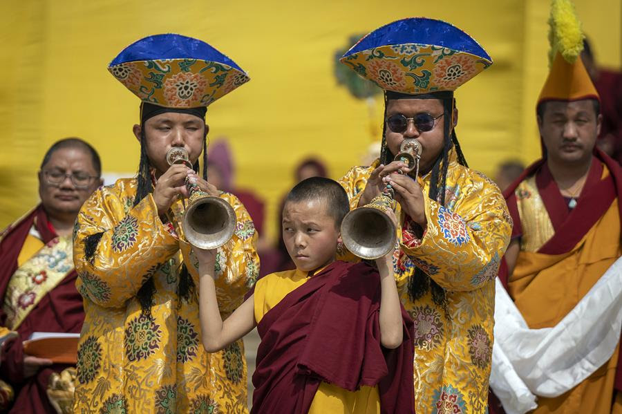 A younger monk standing between two older monks holds their traditional instruments up as they play. All three are wearing traditional clothing. There are other monks in the background.