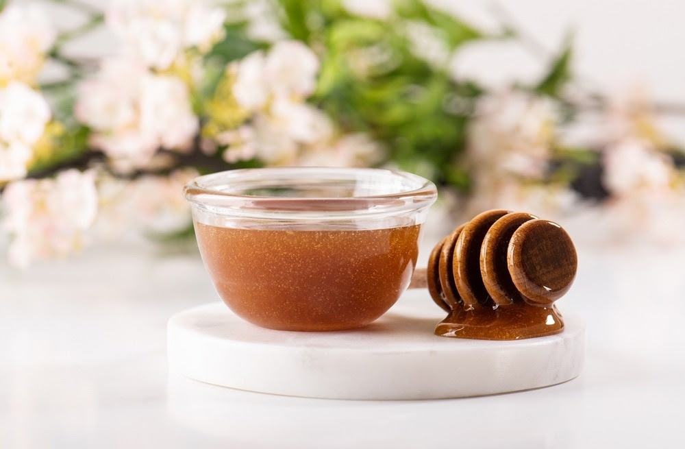 Glass bowl filled with golden honey and a wooden honey dipper resting on a white marble surface, with blurred floral background.