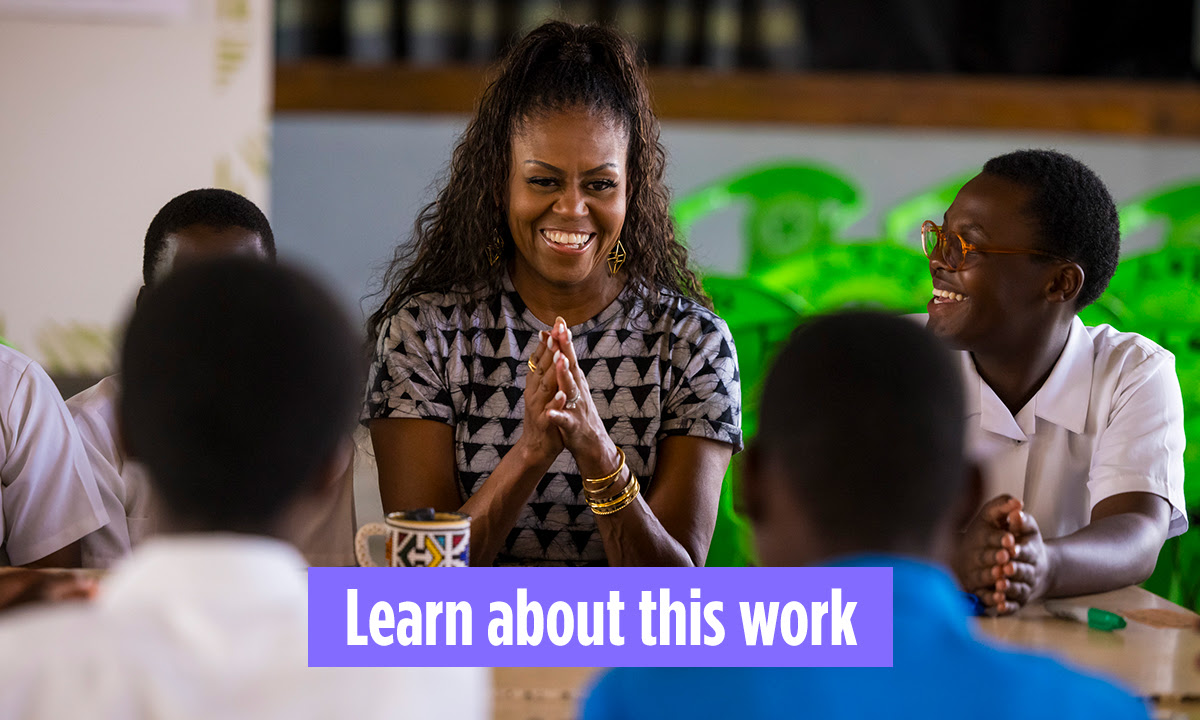 Mrs. Obama is smiling and holding her hands together while sitting at a table surrounded by students with dark skin tones and close cropped hair. At the bottom, a button says "learn about this work"